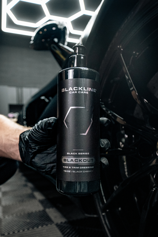 BLACKLINE Blackout Tire & Trim Dressing bottle held by gloved hand beside car under lights on Tampa Home & Garden Store