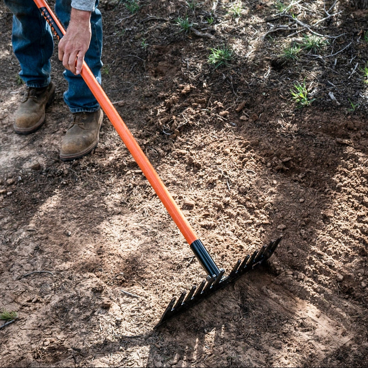 Bert rake by Backslope Tools with orange handle and black tines, held by a worker on dirt on Tampa Home & Garden Store