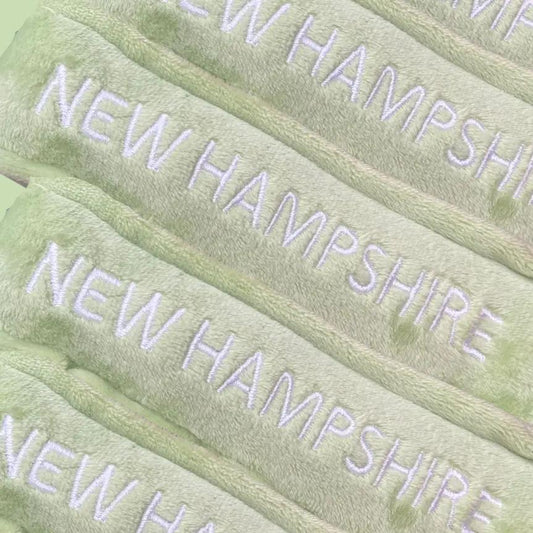 New Hampshire state plush doll on pale green fabric with repeated NEW HAMPSHIRE embroidery on Tampa Home & Garden Store