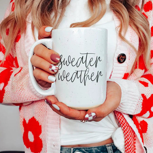 Ceramic mug with white speckled glaze and Sweater Weather script, held by hands in a cardigan on Tampa Home & Garden Store