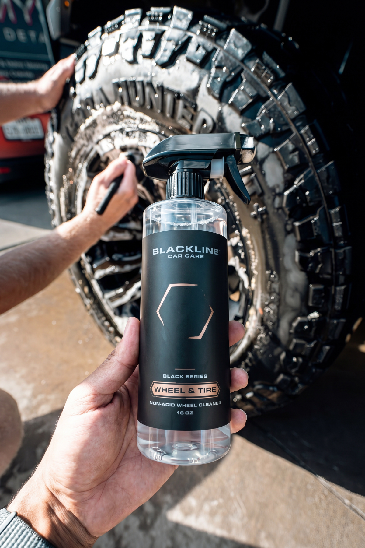 BLACKLINE Wheel N' Tire Cleaner black bottle held before a muddy automotive wheel and tire on Tampa Home & Garden Store
