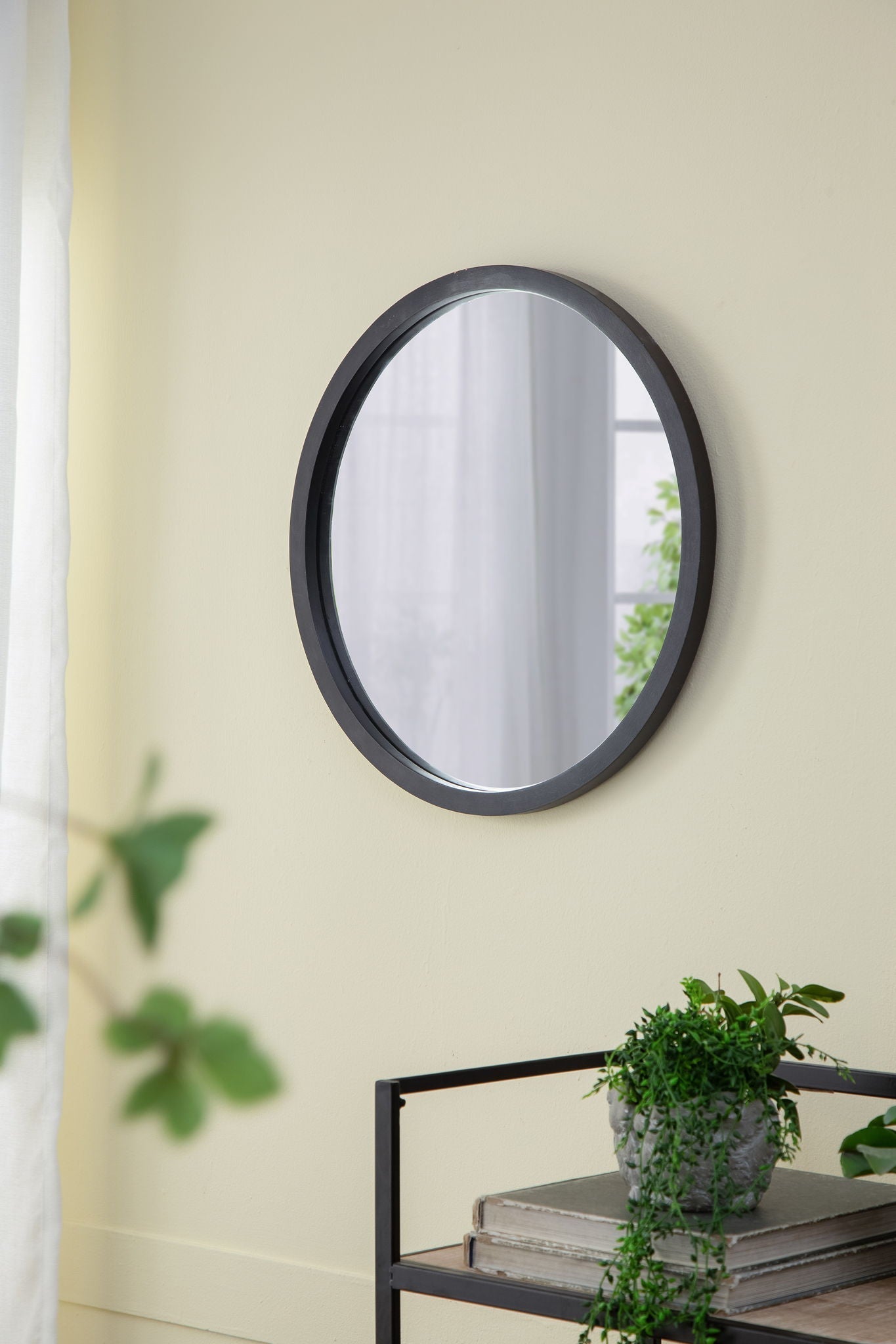 Circle wall mirror with dark wooden frame on pale yellow wall reflects window beside plants on Tampa Home & Garden Store