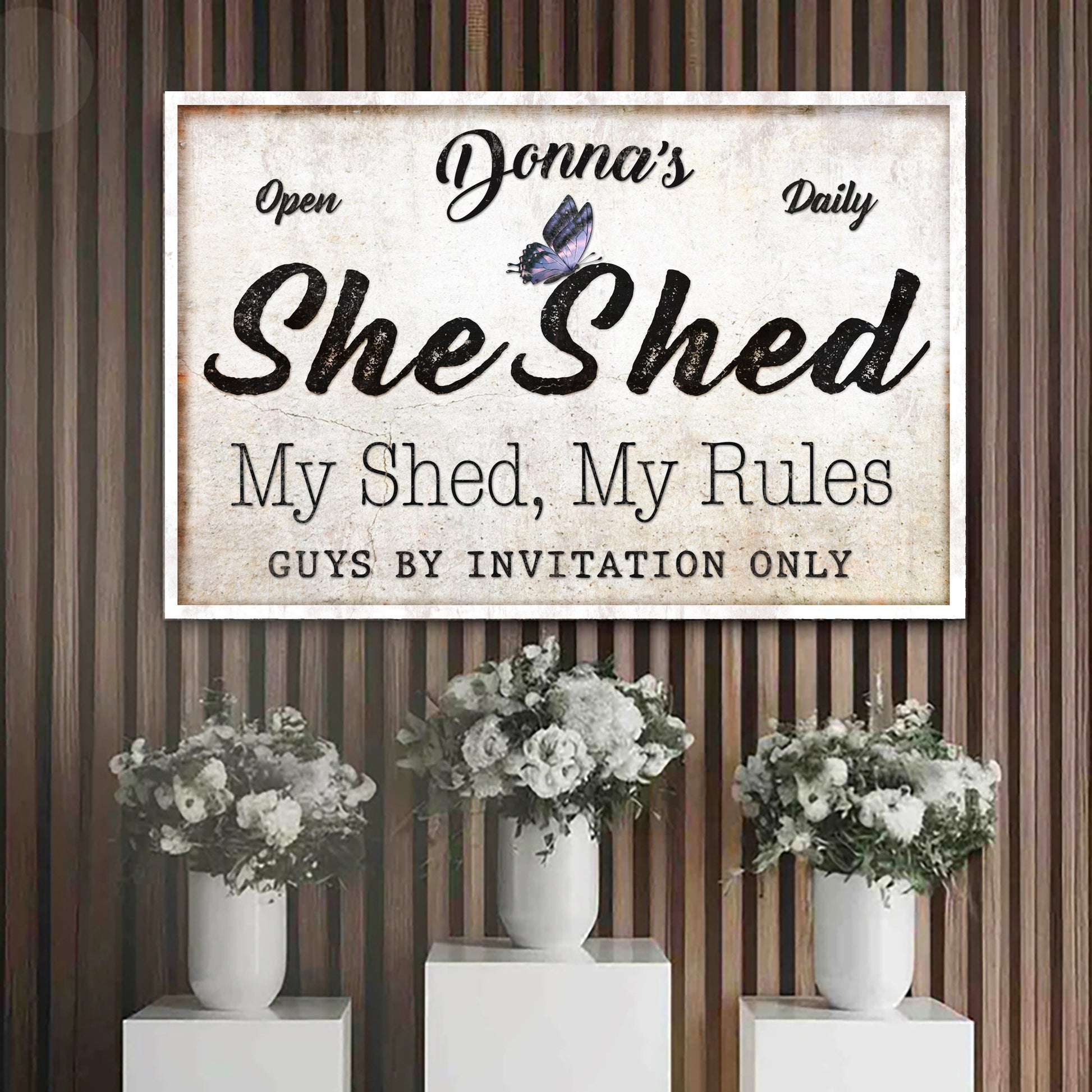 Canvas sign with large She Shed script and butterfly on wood backdrop with white flowers on Tampa Home & Garden Store