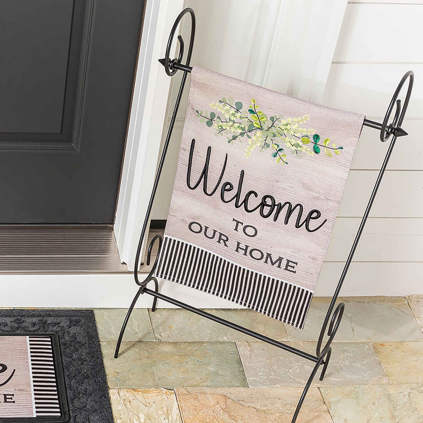 Two wood-grain burlap welcome garden flags on a black metal stand with green leafy design on Tampa Home & Garden Store