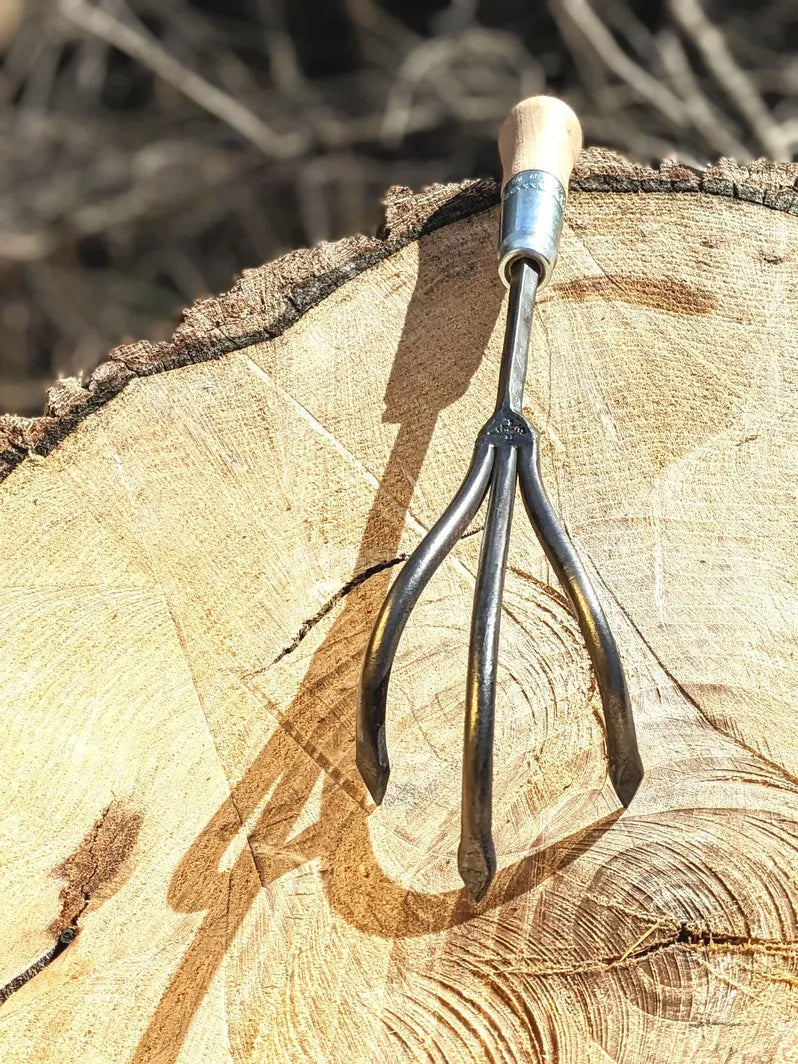 Homestead Iron 3-tine hand-forged cultivator with wooden handle and curved tines on a log stump on Tampa Home & Garden Store