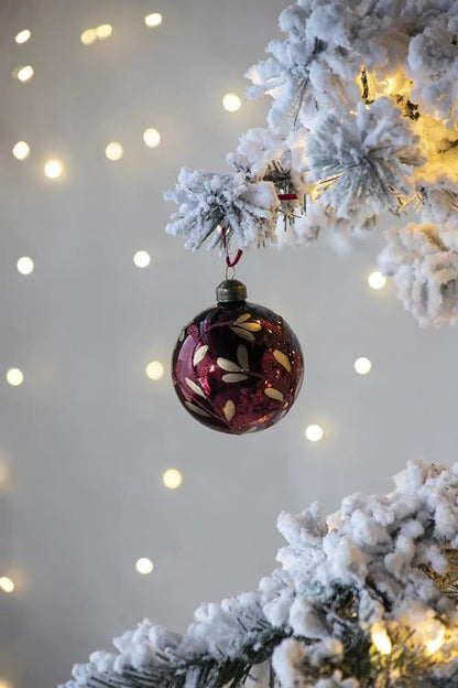 Glass burgundy Christmas ornament ball with gold leaf accents hanging from snow-covered pine on Tampa Home & Garden Store