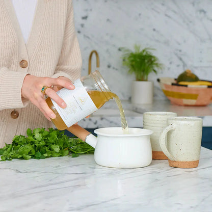 The OWL Reset Organic Cleanse Program broth bottle pours into a white pot with parsley on Tampa Home & Garden Store