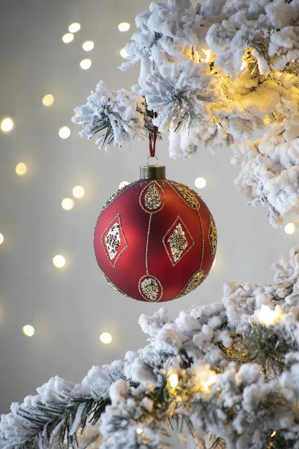 Red glass Christmas ball ornament with gold glitter motifs on snow-dusted tree branch with lights on Tampa Home & Garden Store