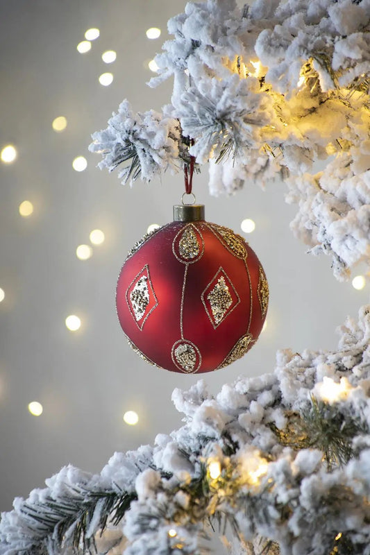 Red glass Christmas ball ornament with gold glitter motifs on snow-dusted tree branch with lights on Tampa Home & Garden Store