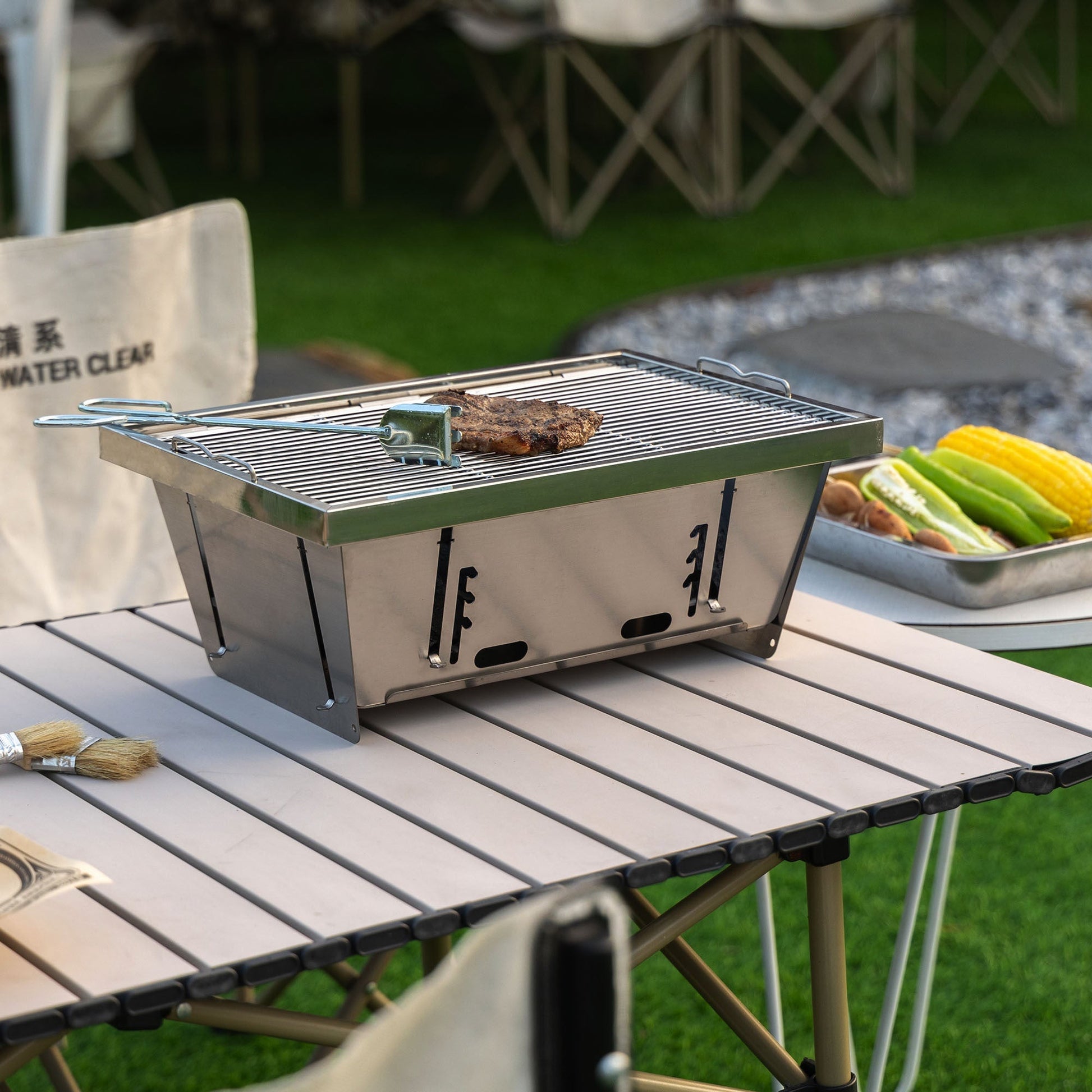 Folded portable BBQ outdoor charcoal grill set in stainless steel with tongs on a white table on Tampa Home & Garden Store