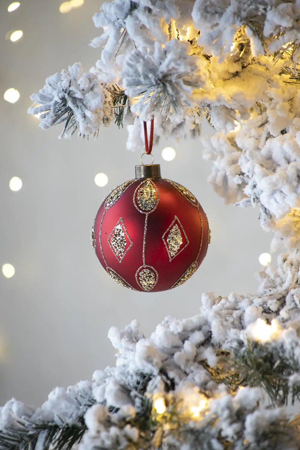 Red Christmas ball ornament with gold glitter accents hanging from a snow-dusted pine branch on Tampa Home & Garden Store