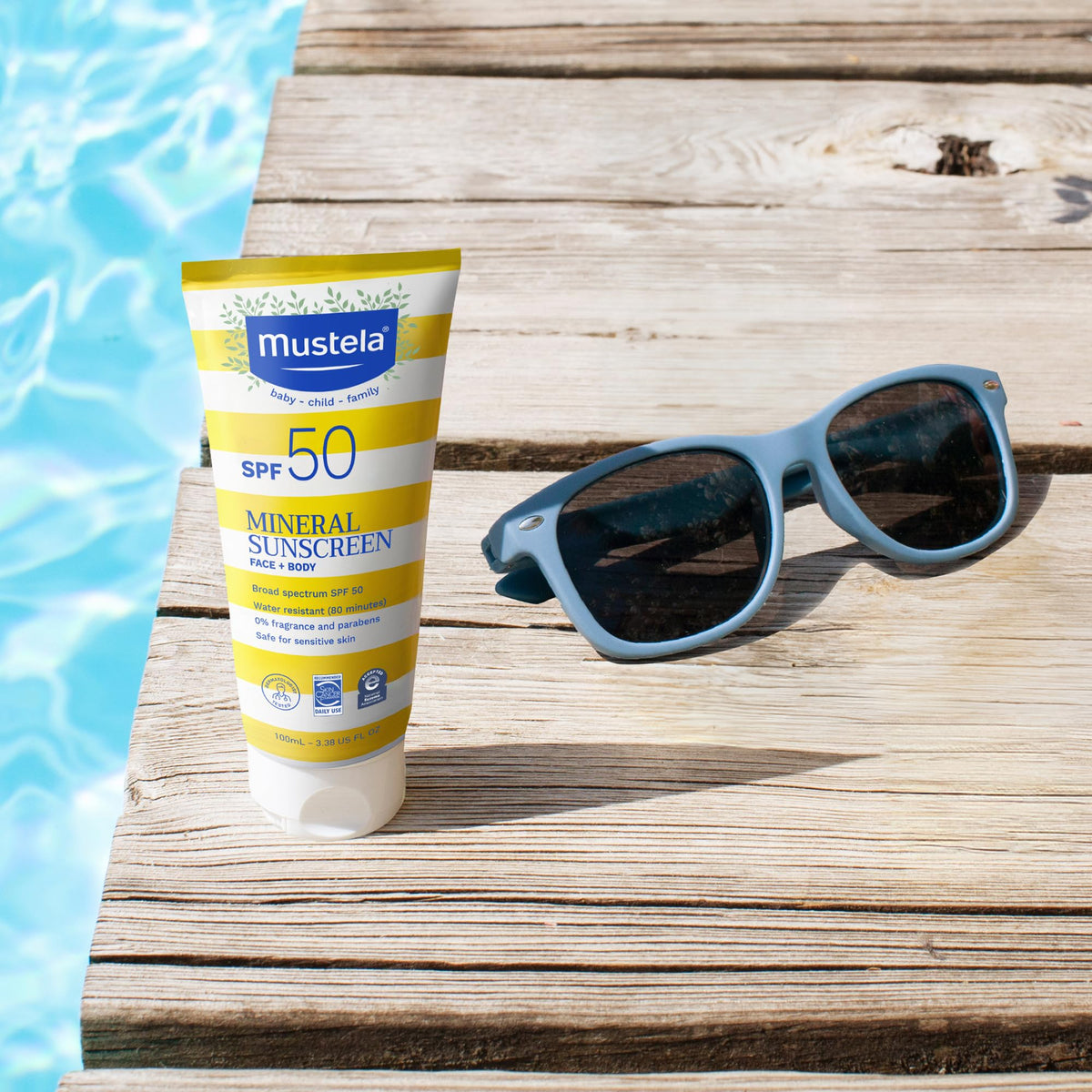 Mustela SPF 50 sunscreen tube with yellow stripes beside blue sunglasses on a wooden deck on Tampa Home & Garden Store