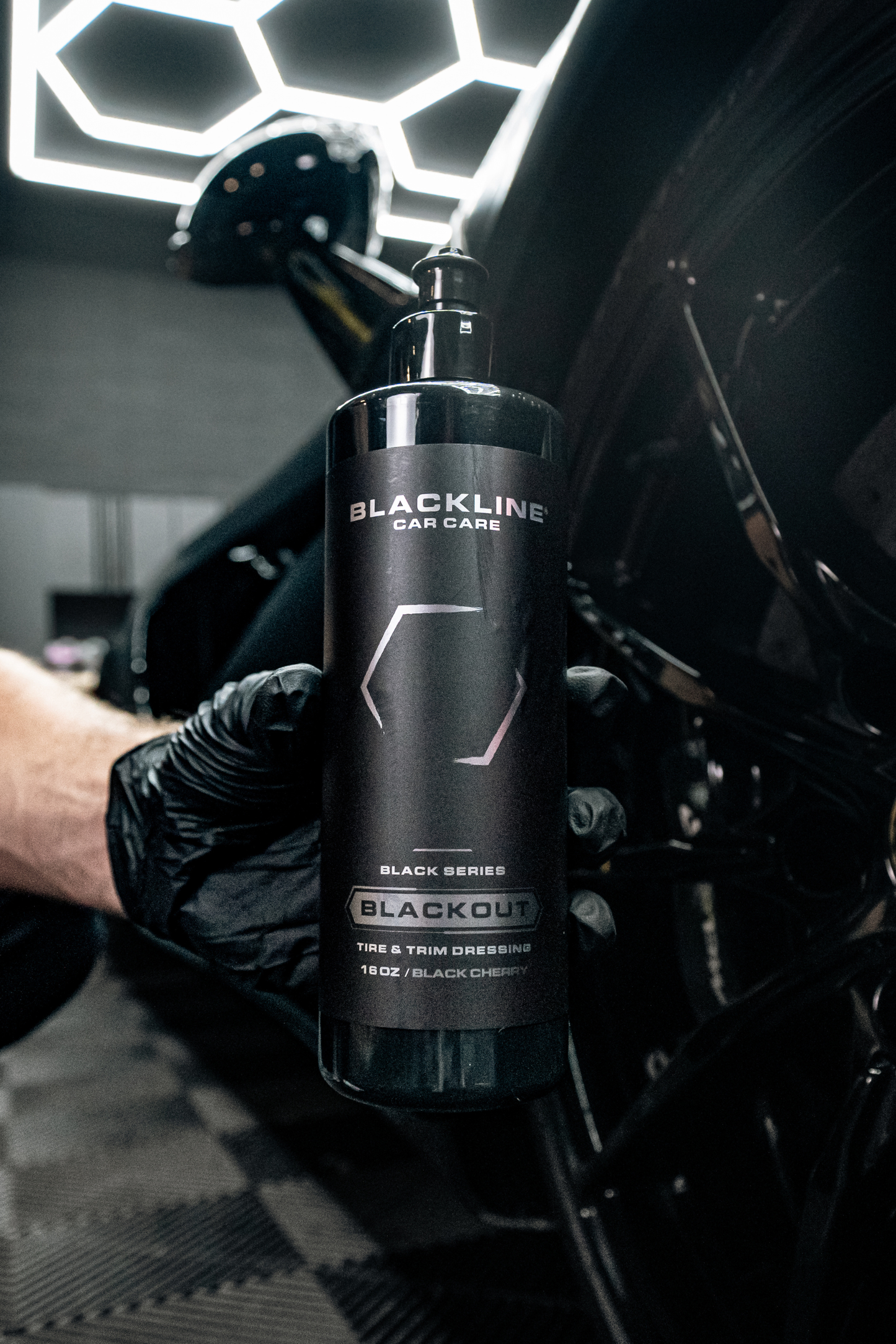 BLACKLINE Blackout Tire & Trim Dressing bottle held by gloved hand beside car under lights on Tampa Home & Garden Store