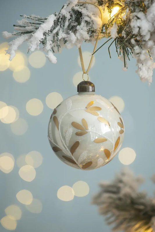 White glass Christmas ball ornament with gold leaf accents, hanging from snow-dusted pine on Tampa Home & Garden Store