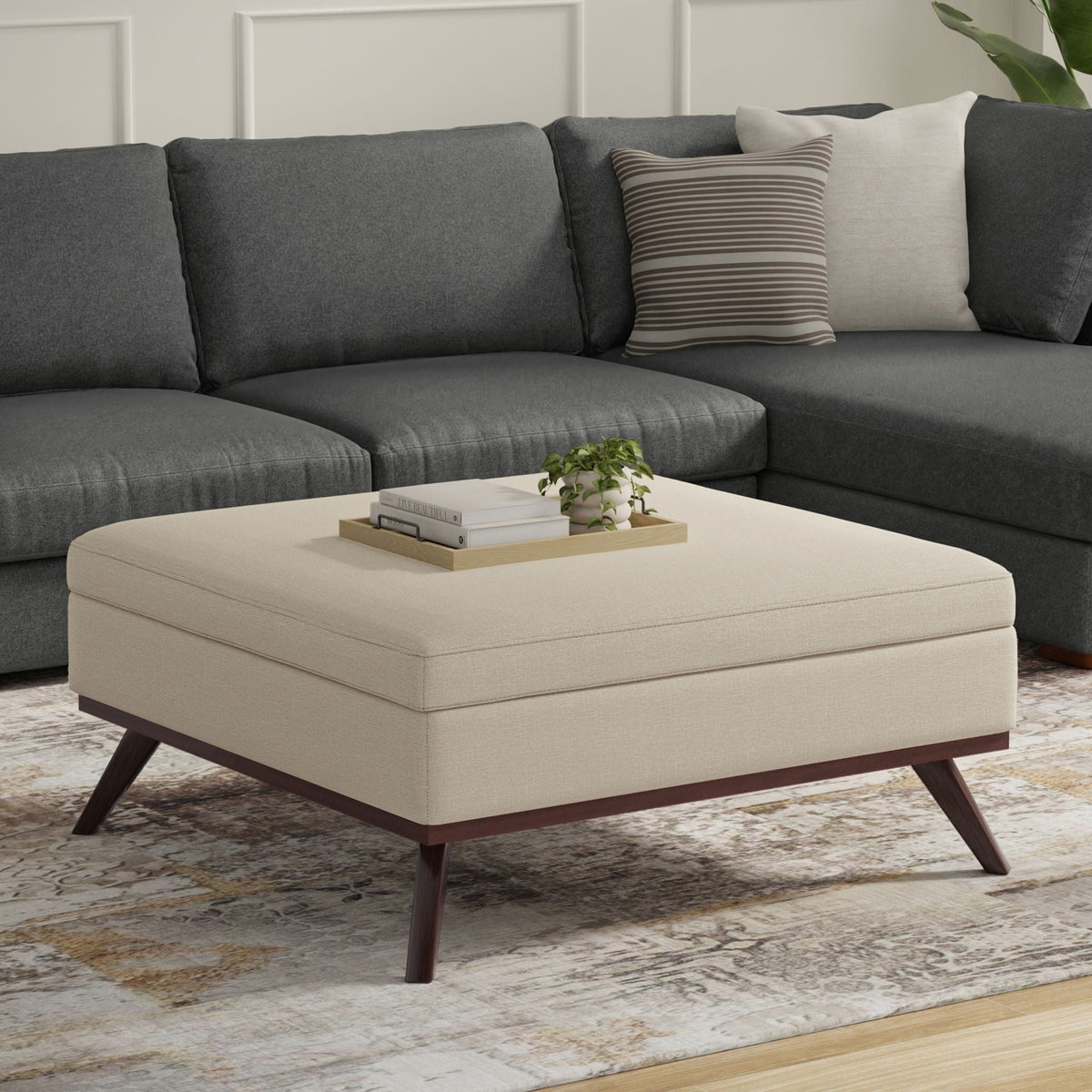 Beige fabric storage ottoman with lift-top, wooden legs, tray of books and a plant on top on Tampa Home & Garden Store