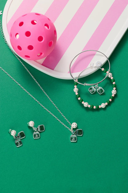 Barbie™ Pickleball Charm Bangle by ALEX AND ANI, silver hoop bracelet with pink and green beads and pickleball charms beside a pink perforated ball on a striped plate on Tampa Home & Garden Store