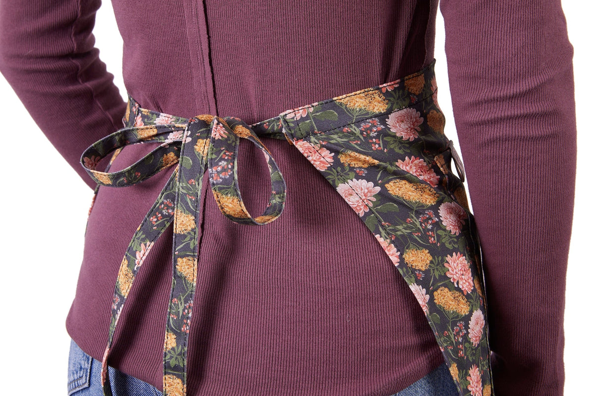 Gardener's Half Apron with floral belt tied in a bow around a purple rib-knit top on Tampa Home & Garden Store