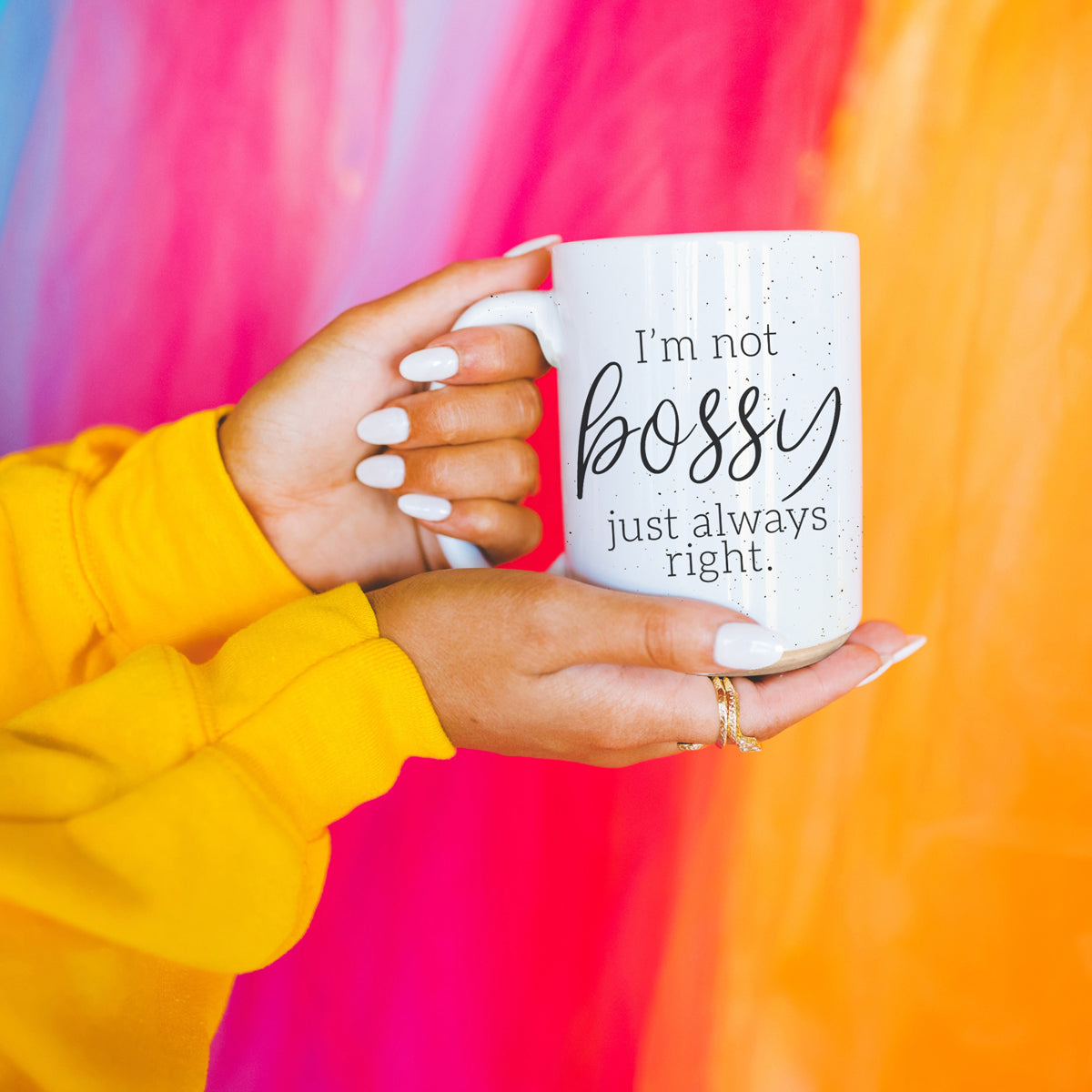 Bossy 16oz coffee mug by Gia Roma, white speckled ceramic held in yellow-sleeved hands against a vibrant backdrop on Tampa Home & Garden Store