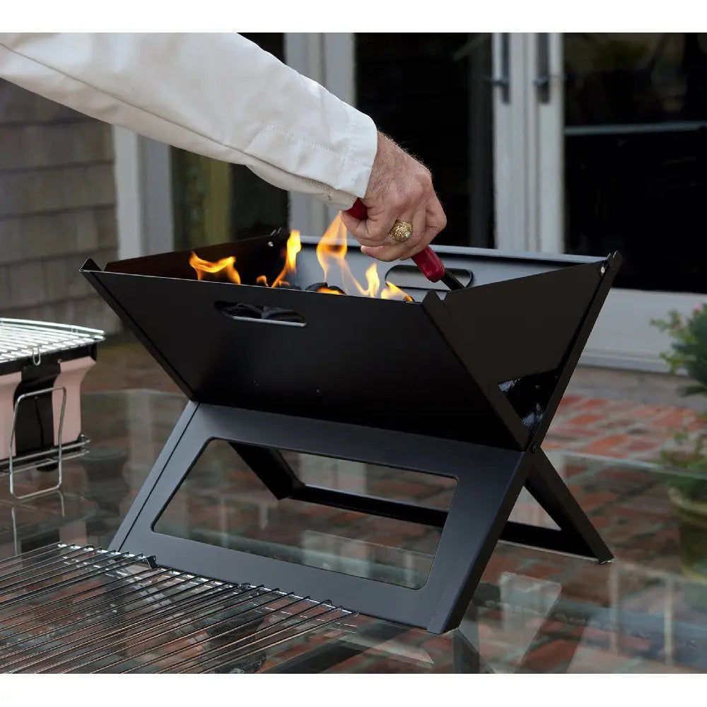 Black Notebook Charcoal Grill with folding X-frame; hand tending orange flames on patio on Tampa Home & Garden Store