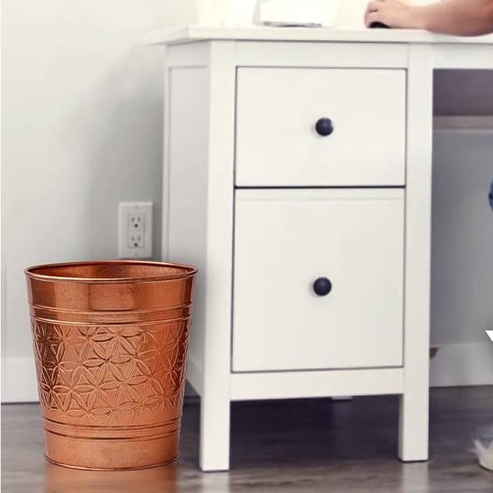 Copper finish metal trash can waste basket with embossed geometric pattern beside a white two-drawer cabinet on Tampa Home & Garden Store