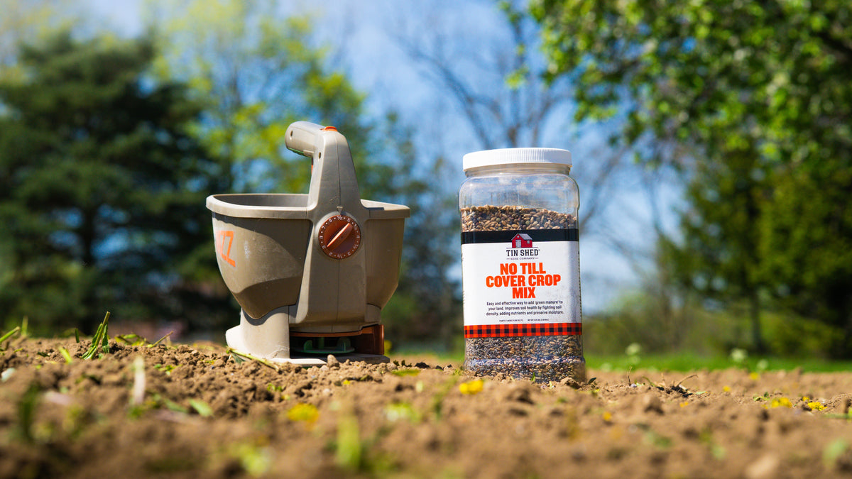 Tin Shed Seed Company No Till Cover Crop Mix seeds in a clear jar beside a garden spreader on Tampa Home & Garden Store