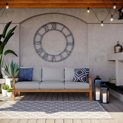 Marina Outdoor Teak Sofa with gray cushions in a modern patio setup with a large clock backdrop on Tampa Home & Garden Store