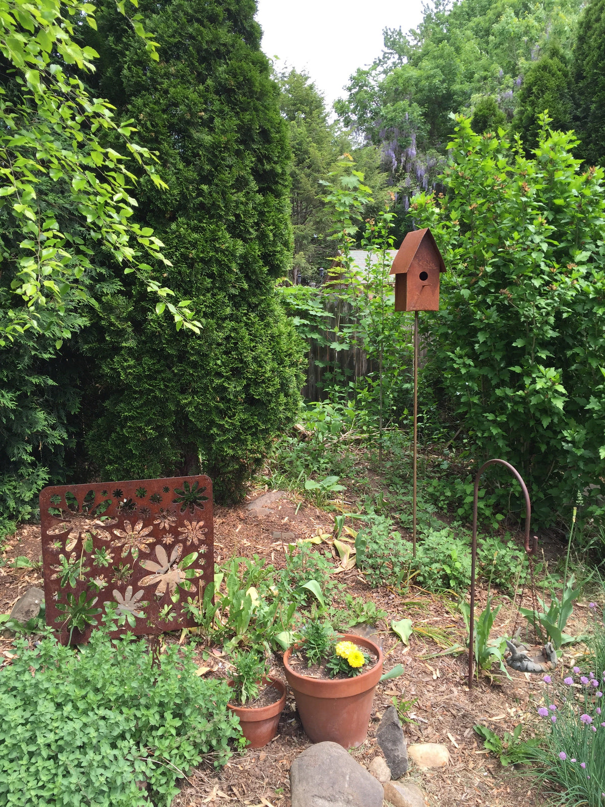 Rust-colored metal garden art panel with floral cutouts and a matching birdhouse on a stake in a garden on Tampa Home & Garden Store