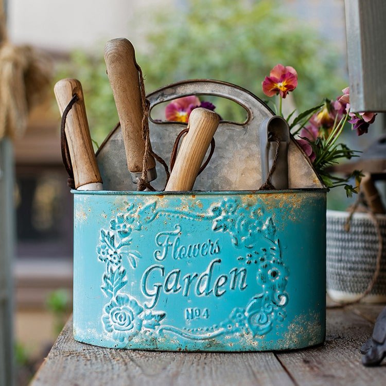 Blue metal basket tool organizer with wooden-handled gardening tools and pink flowers on Tampa Home & Garden Store