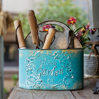Blue metal basket tool organizer with wooden-handled gardening tools and pink flowers on Tampa Home & Garden Store