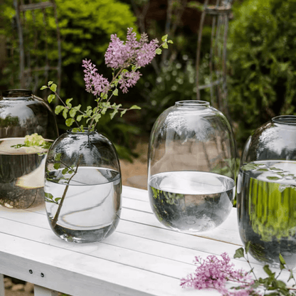 Modern clear glass vase trio on a white outdoor table with water, stems, garden backdrop on Tampa Home & Garden Store