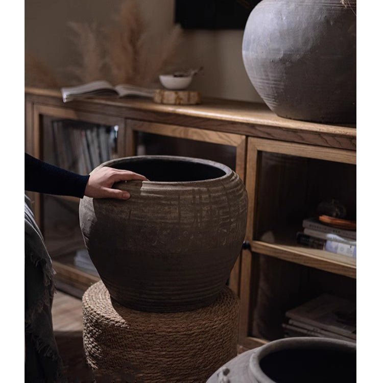 Traditional Round Belly Zen Style Ceramic Vase in charcoal glaze; hand on rim atop a woven stool on Tampa Home & Garden Store