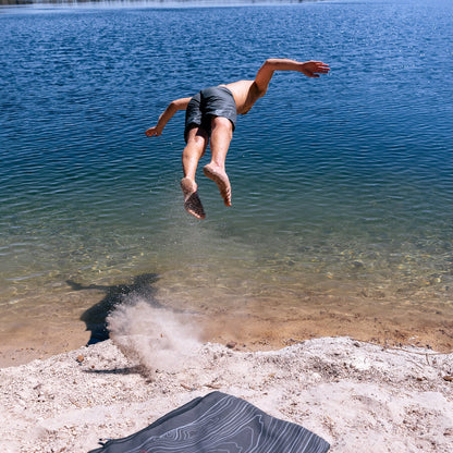 Terrain Towel by Laek, dark striped beach towel on sandy shore beside clear blue water on Tampa Home & Garden Store