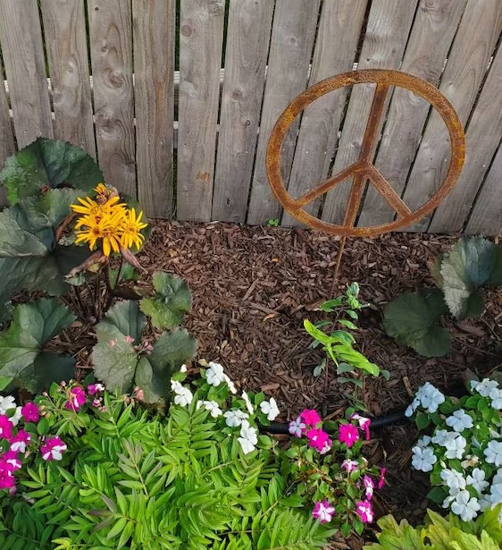 Outdoor peace sign metal sculpture in rust finish leaning against a fence among blooms on Tampa Home & Garden Store