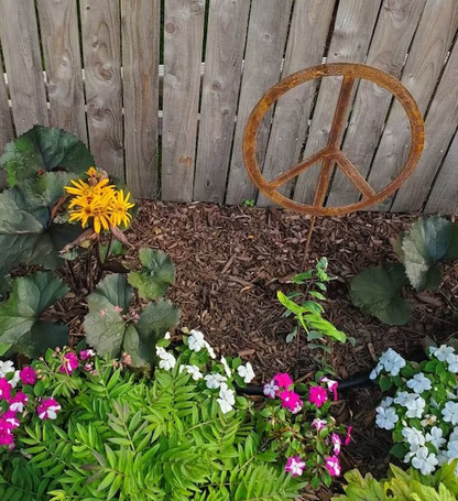 Outdoor peace sign metal sculpture in rust finish leaning against a fence among blooms on Tampa Home & Garden Store