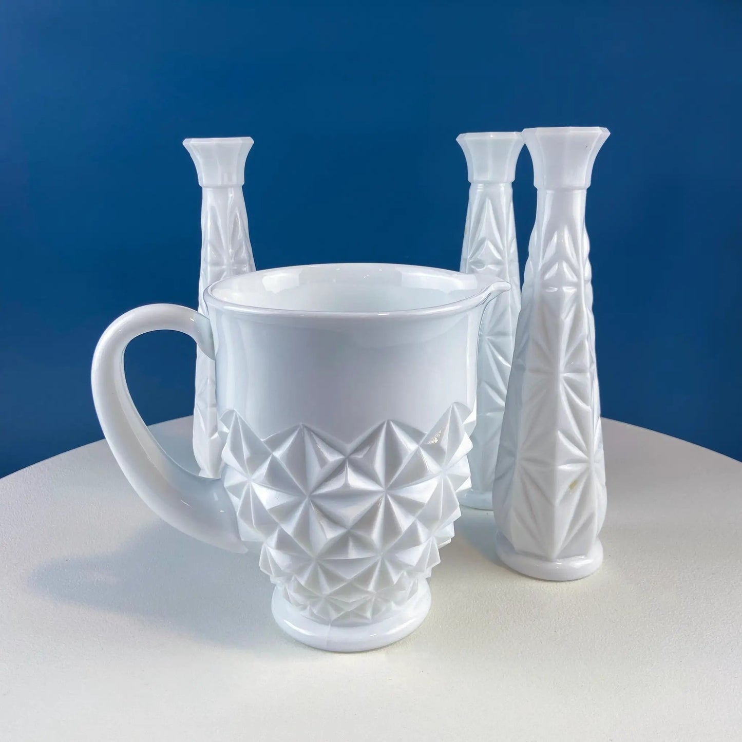 Milk glass pitcher with geometric facets surrounded by four candlesticks on a blue backdrop on Tampa Home & Garden Store