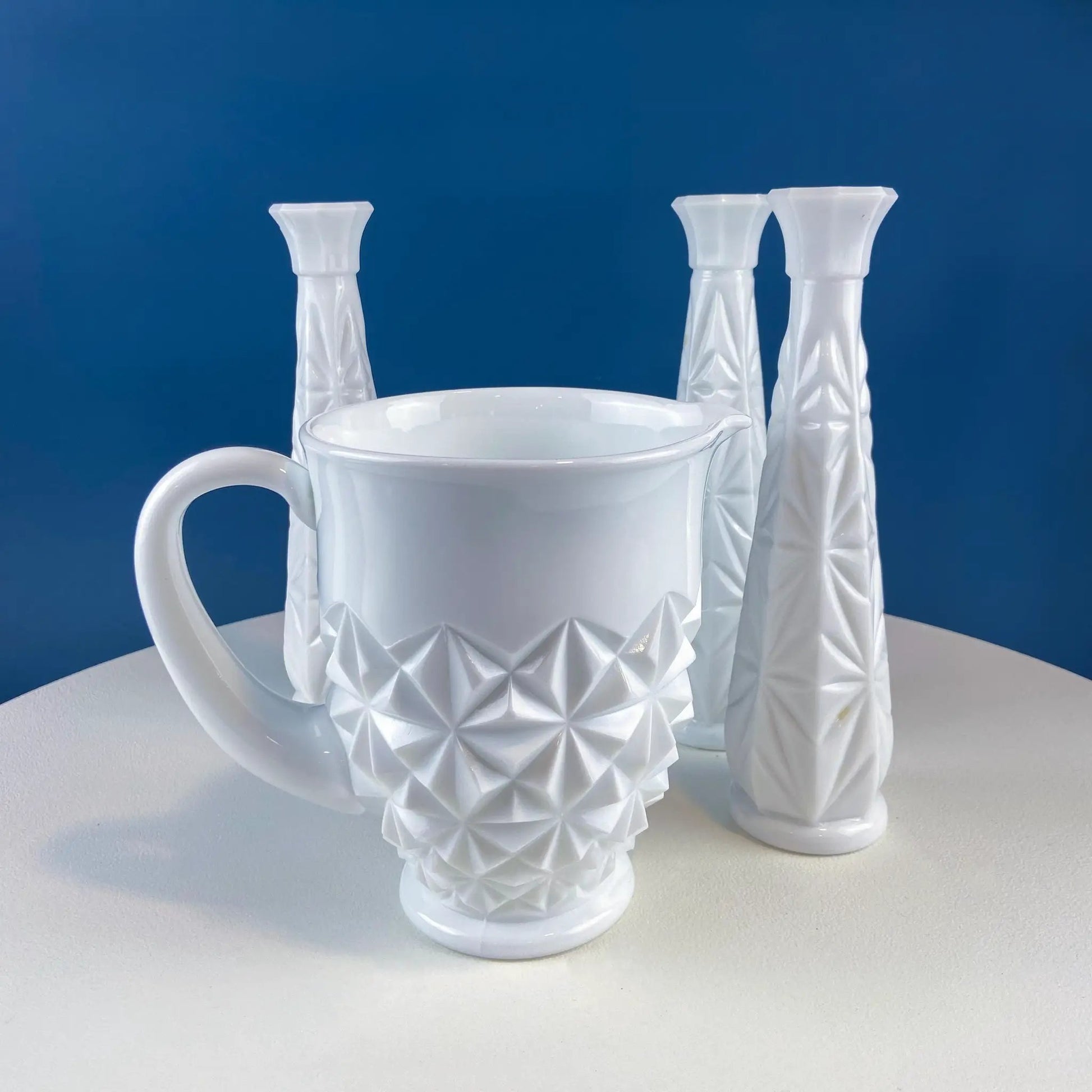 Milk glass pitcher with geometric facets surrounded by four candlesticks on a blue backdrop on Tampa Home & Garden Store