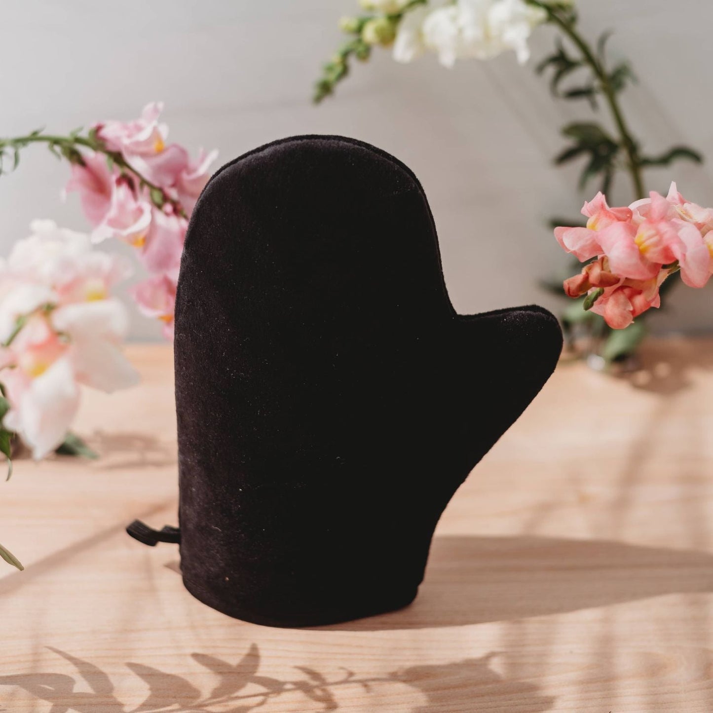 Black velvet self-tanning mitt on a wooden table with pink blossoms in the background on Tampa Home & Garden Store