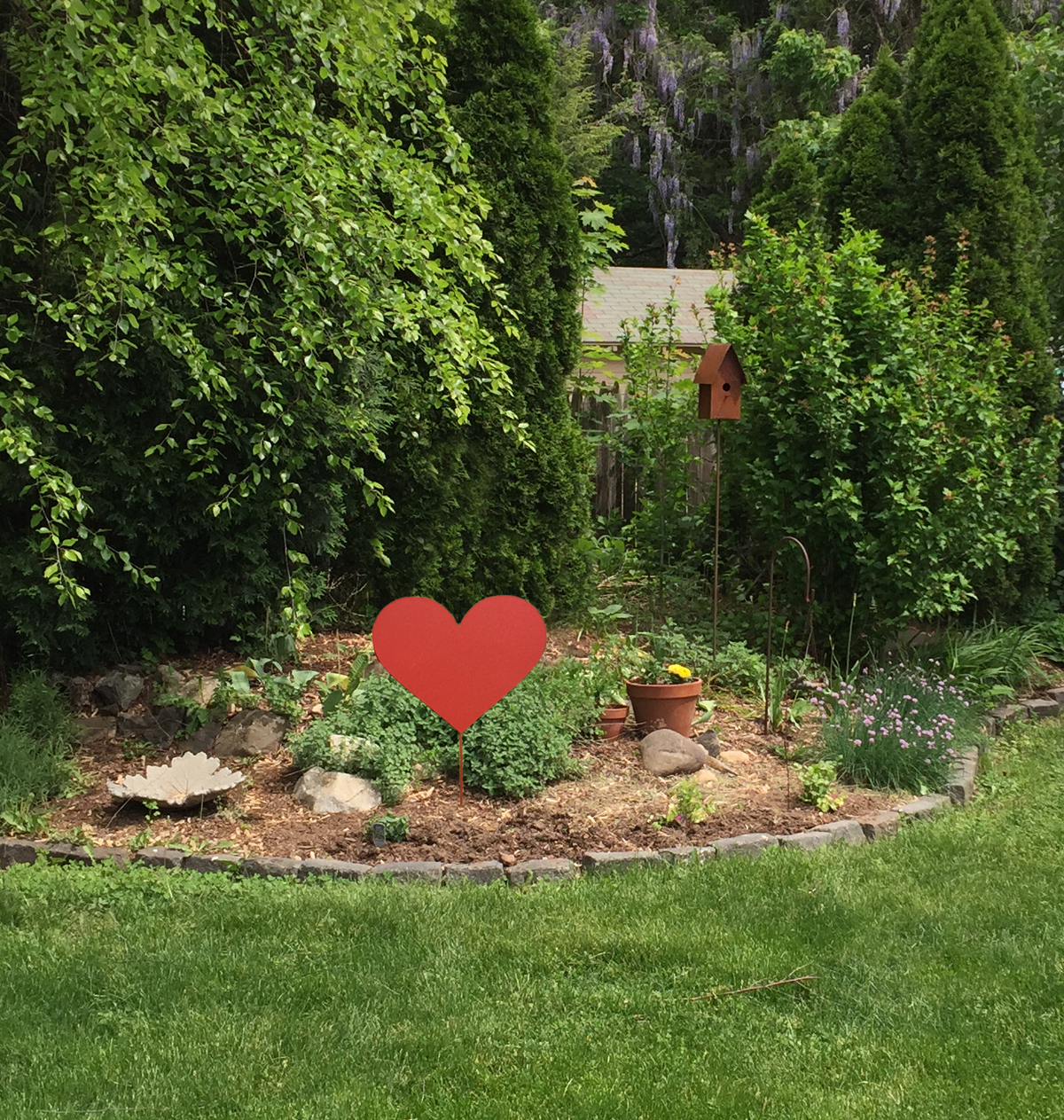 Red metal heart garden stake by Rustica Ornamentals in a rock-bordered garden with potted plants on Tampa Home & Garden Store