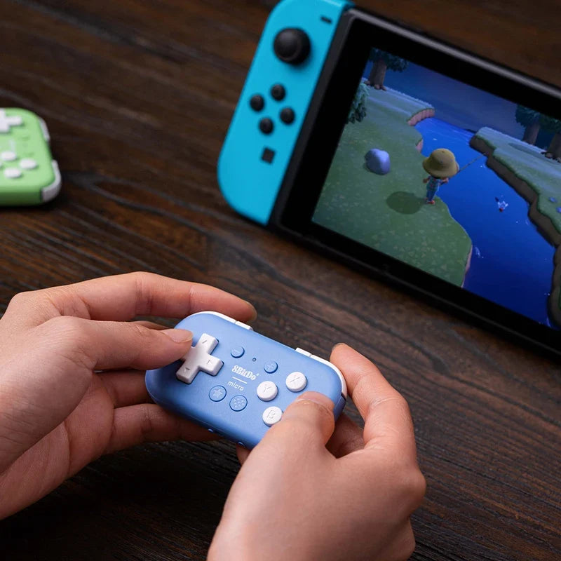 Blue 8BitDo micro Bluetooth gamepad held in hands; Nintendo Switch in background displaying a game on Tampa Home & Garden Store