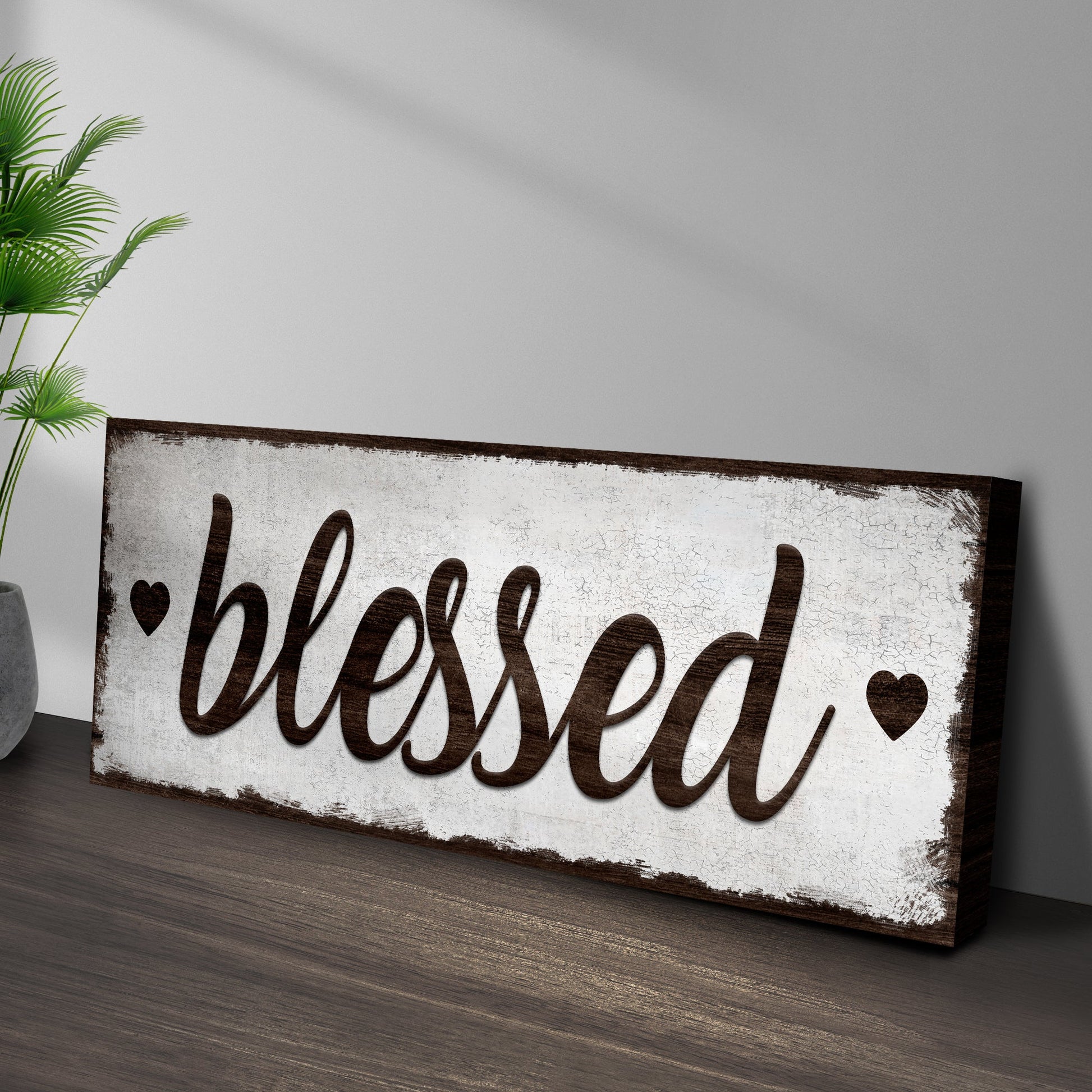 Blessed Sign canvas with distressed white panel and dark brown cursive lettering, shown beside a green palm plant on Tampa Home & Garden Store