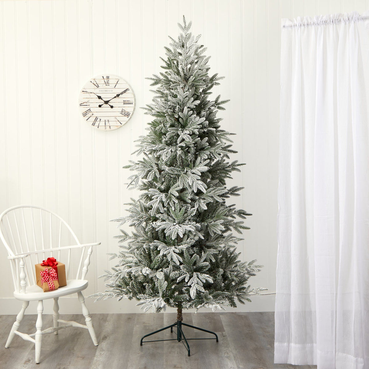 7.5 ft flocked Manchester Spruce tree with snow-dusted branches indoors by white curtain on Tampa Home & Garden Store