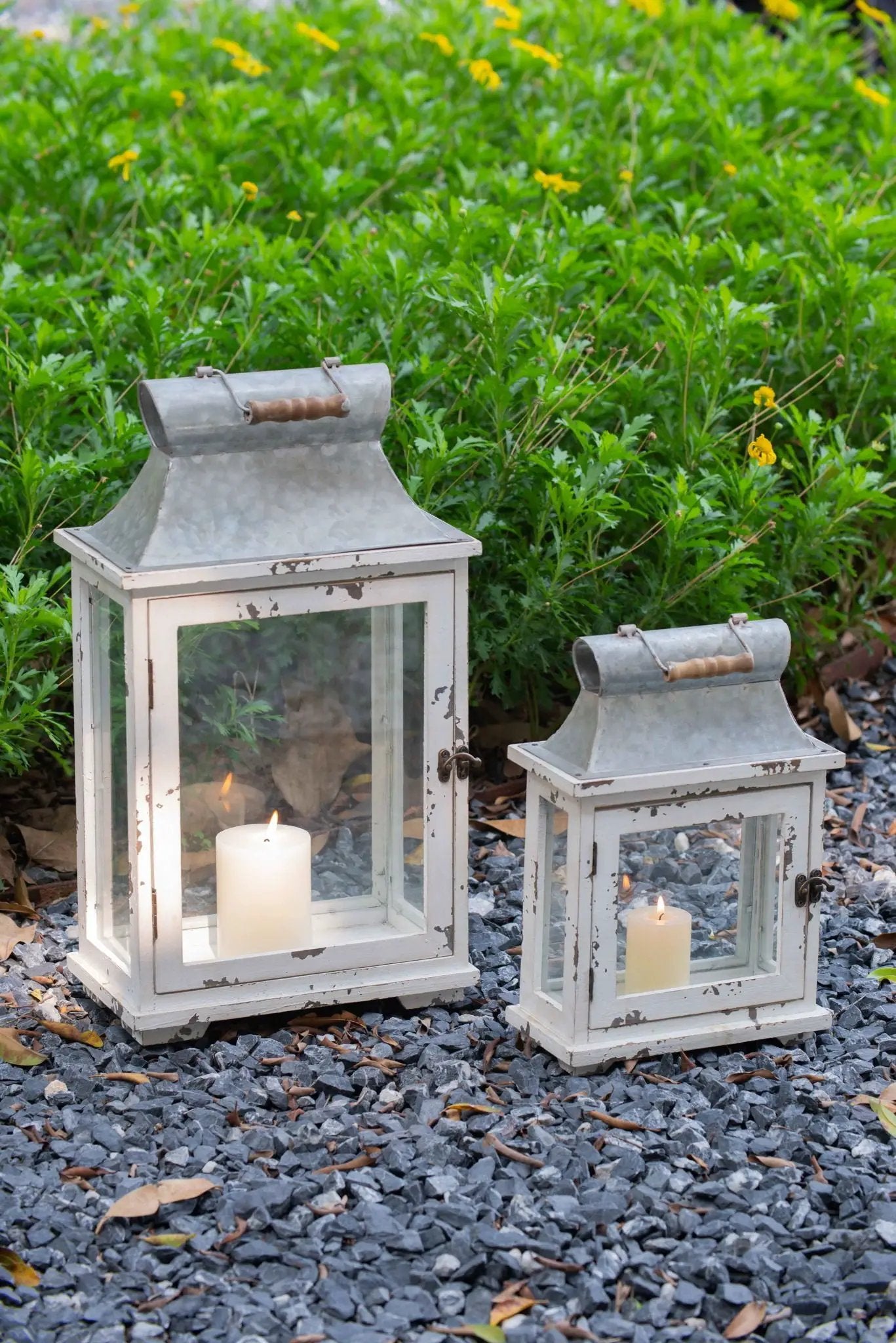 Two white distressed wooden lanterns with metal tops, glass panes and lit candles in garden on Tampa Home & Garden Store