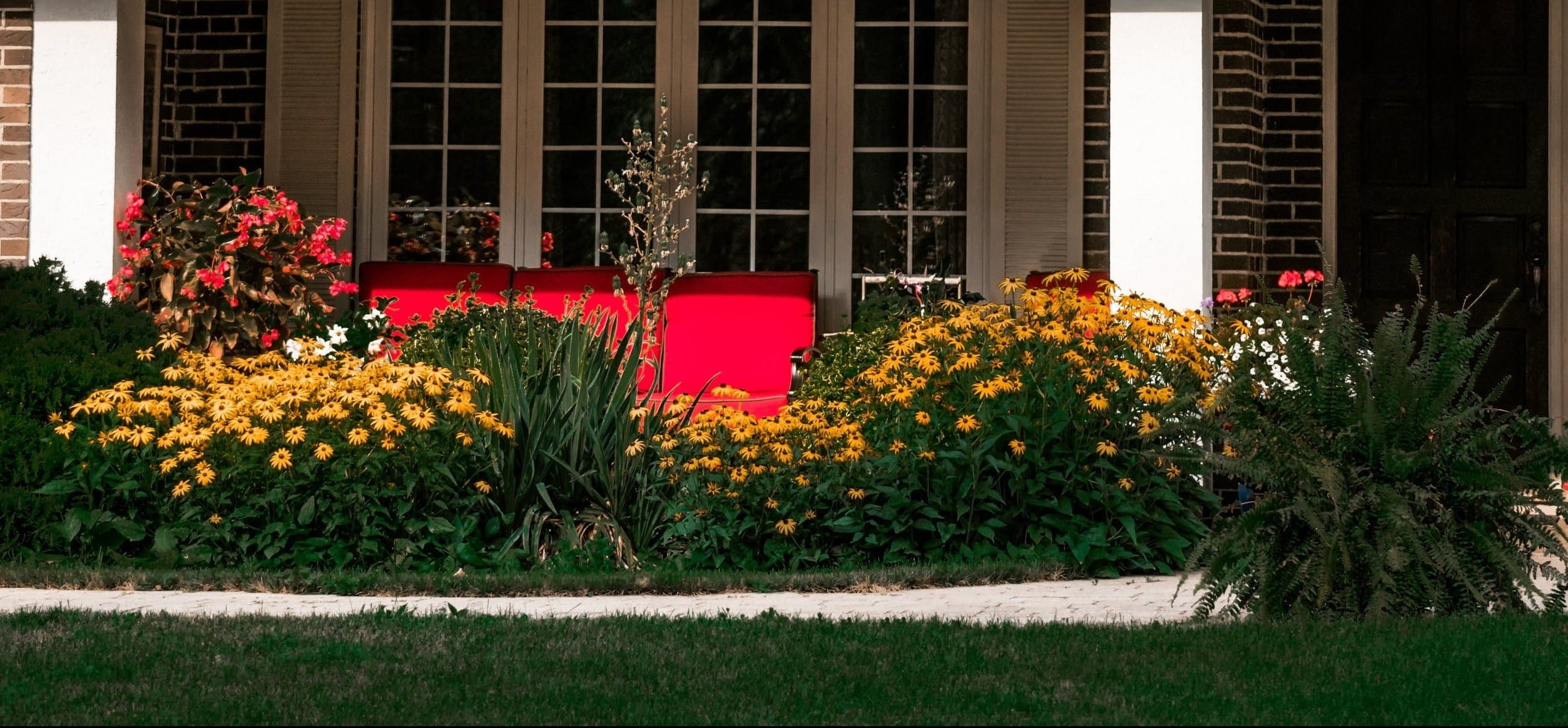 A brick house facade with large patio, a red outdoor sofa, and a landscaped garden filled with yellow flowers and green plants on Tampa Home & Garden Store.