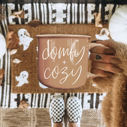 Gia Roma pink coffee mug with white comfy + cozy script, held by a hand with fuzzy sleeve over a patterned rug on Tampa Home & Garden Store