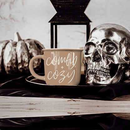 Beige ceramic coffee mug with white script comfy + cozy beside a decorative skull on a tray on Tampa Home & Garden Store