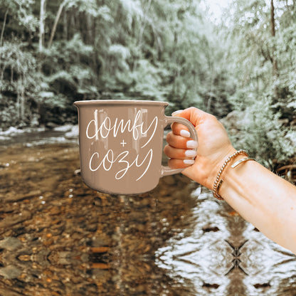 Coffee mug Gia Roma taupe with white comfy + cozy script, held by hand beside a river backdrop on Tampa Home & Garden Store