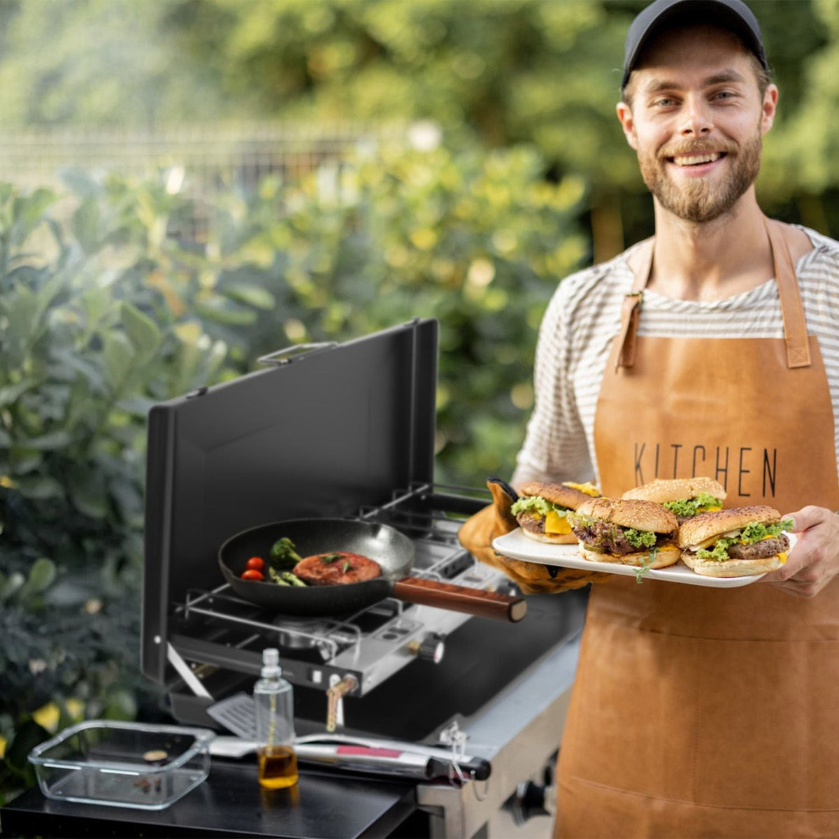 Mercantile Mountain 3-Burner Portable Propane Stove on a black grill; man in brown apron holds burgers on Tampa Home & Garden Store