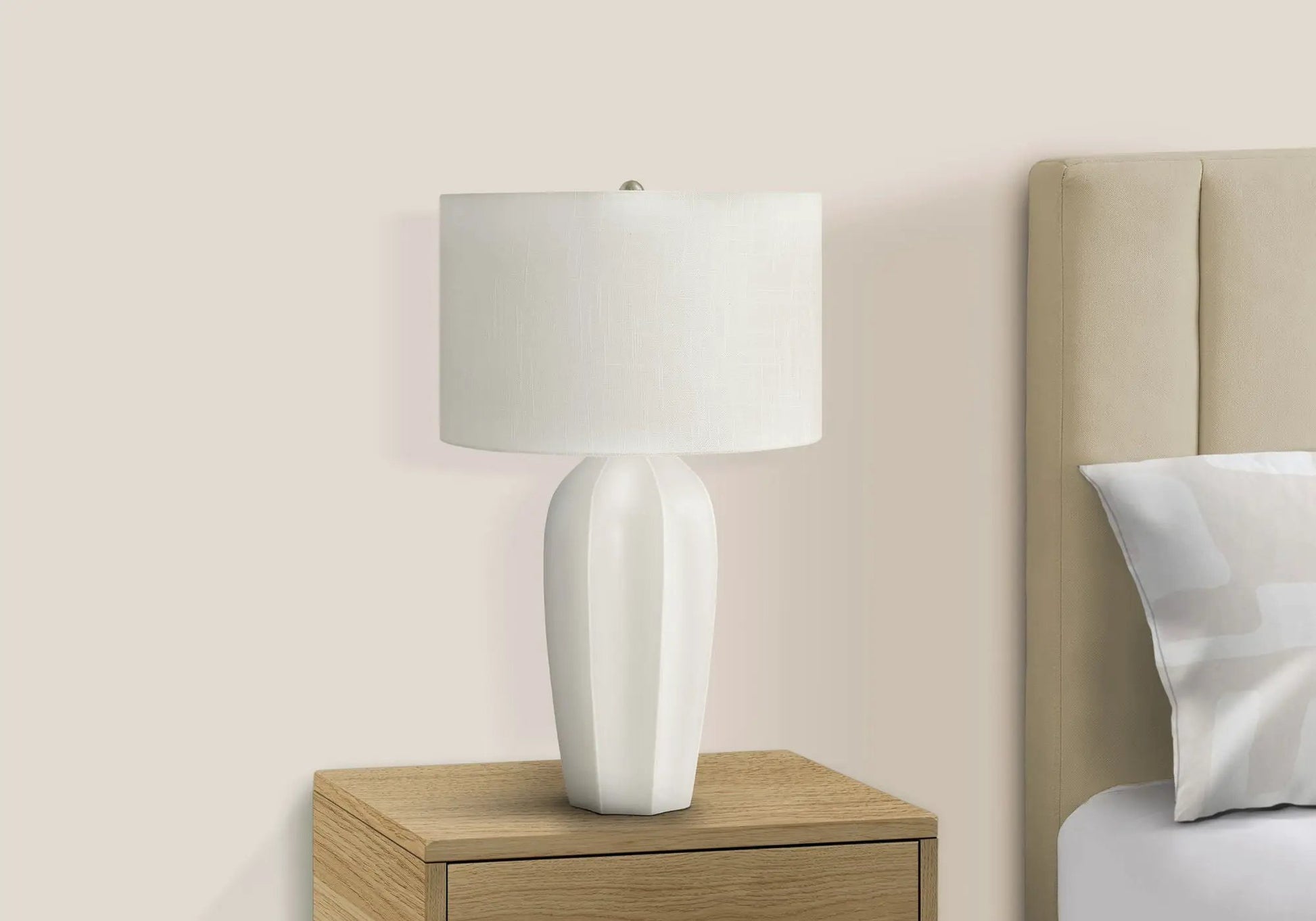 Table lamp with cream ceramic base and white linen shade on a wood nightstand beside a beige bed on Tampa Home & Garden Store