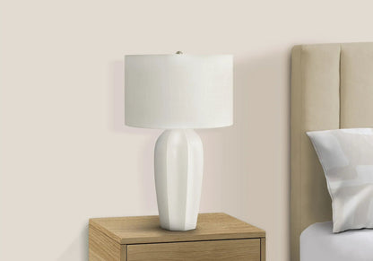 Table lamp with cream ceramic base and white linen shade on a wood nightstand beside a beige bed on Tampa Home & Garden Store