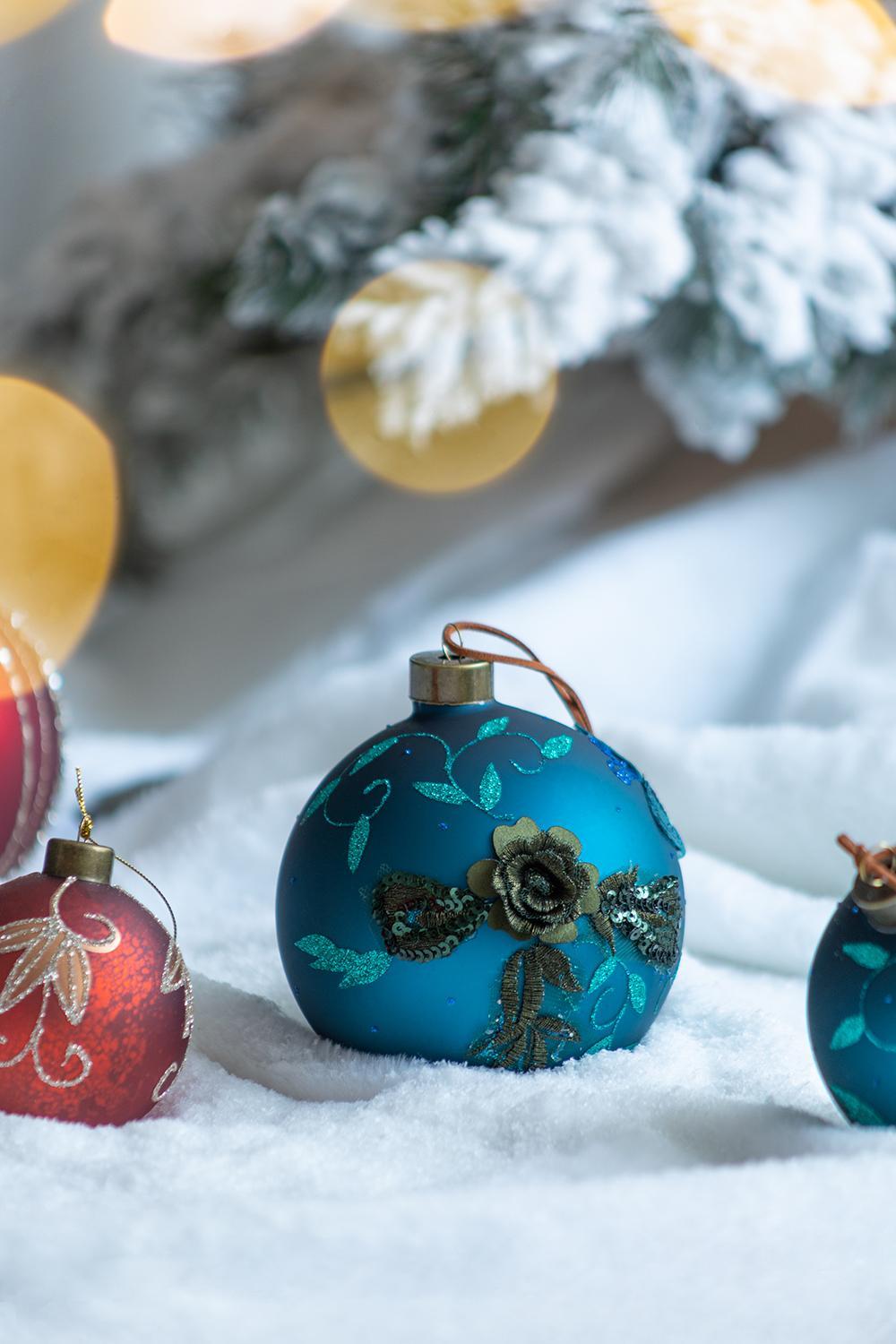 Blue glass ball ornament with teal floral design and gold cap among red ornaments on snow on Tampa Home & Garden Store